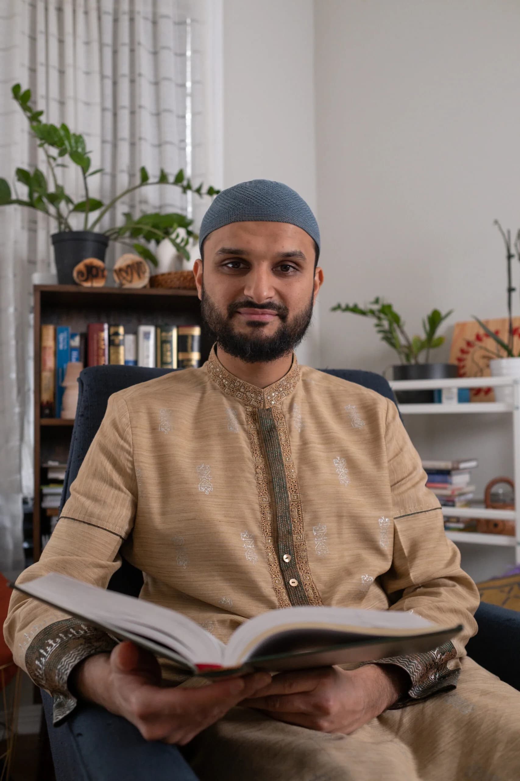 Imam Shuaib seated with an open Qur'an in his study, bookshelves behind him
